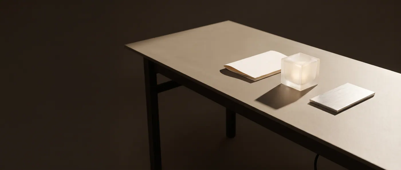 Une table de studio : un carnet ouvert, un cube de verre dépoli et une plaque de métal, sous une lumière d'en haut.